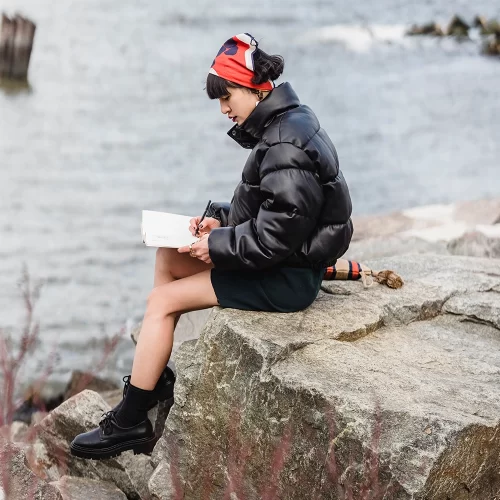 Young woman in a puffy coat sitting on a rock by the water and writing in a notebook