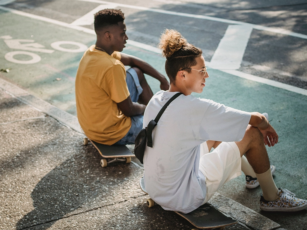 Two young men sitting on their skateboards on the sidewalk