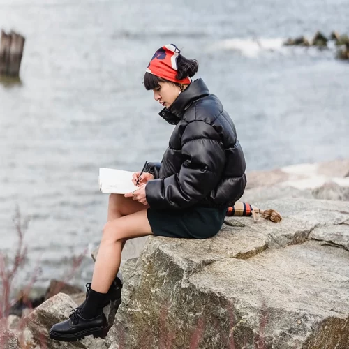 Young woman in a puffy coat sitting on a rock by the water and writing in a notebook